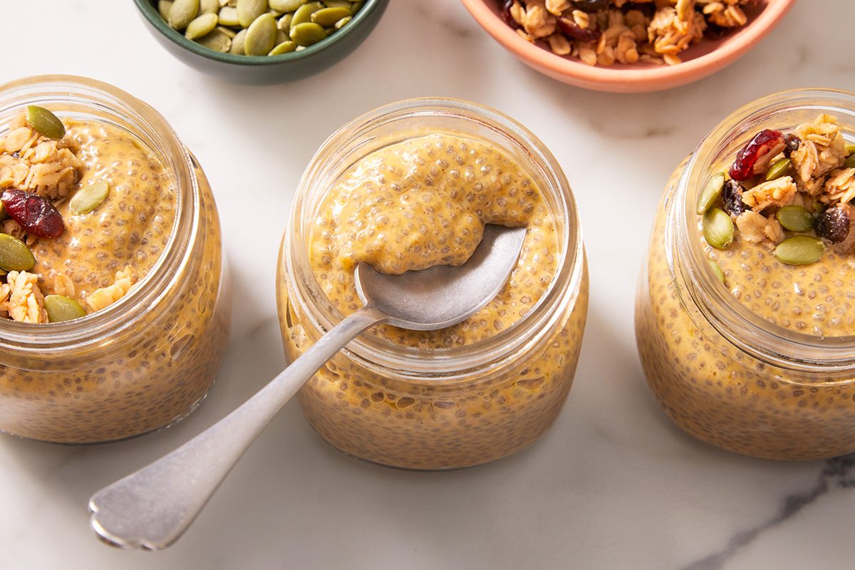 Three glass jars filled with creamy pumpkin chia pudding sit on a white surface, one with a spoon inside. Bowls of granola and pumpkin seeds are visible in the background.