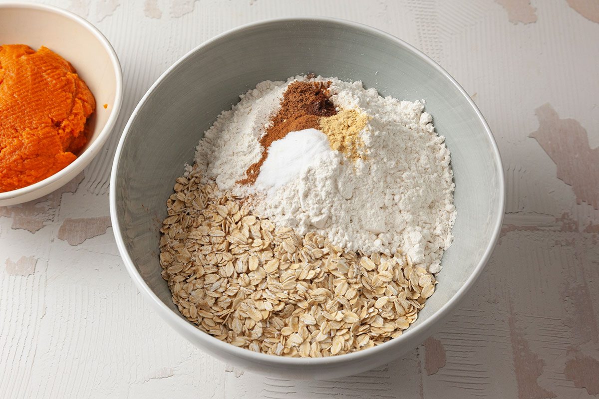 Overhead shot of a mixing bowl with dry ingredients rolled oats, flour, baking soda, ground ginger, and cinnamon on a light surface; a separate bowl of orange pumpkin purée is partially visible beside it;