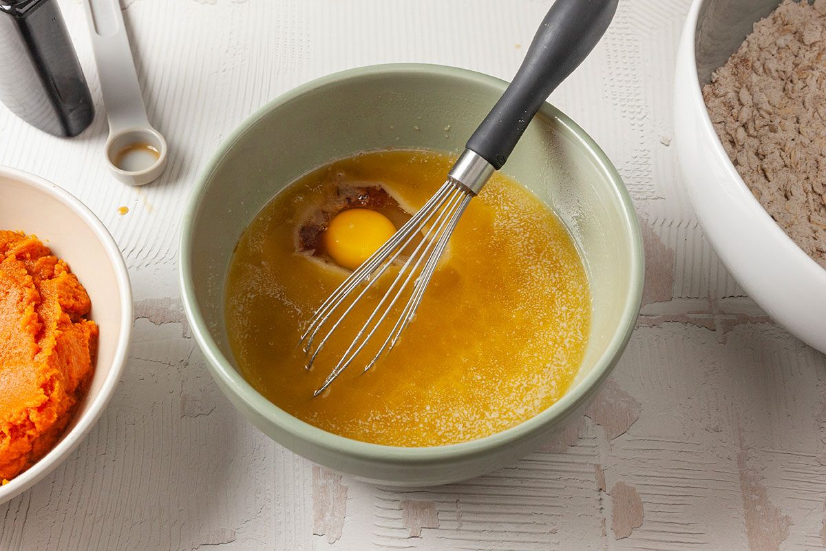 Overhead shot of a mixing bowl with a whisk, containing melted butter, an egg, and vanilla extract; nearby sit a bowl of flour and a bowl of orange pumpkin purée on a light surface.
