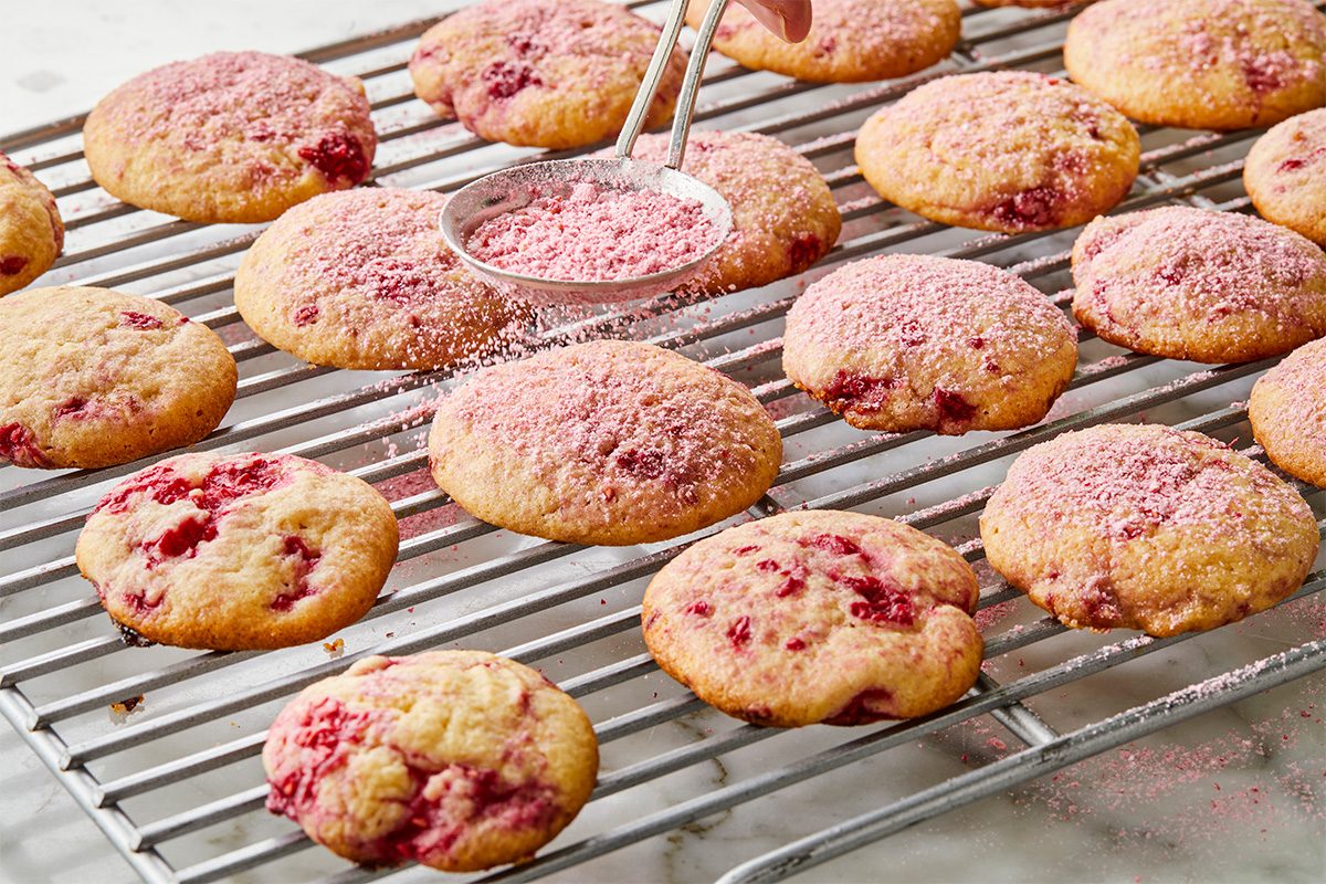 A hand sprinkles pink powdered sugar over freshly baked cookies with red berry pieces, cooling on a metal wire rack.