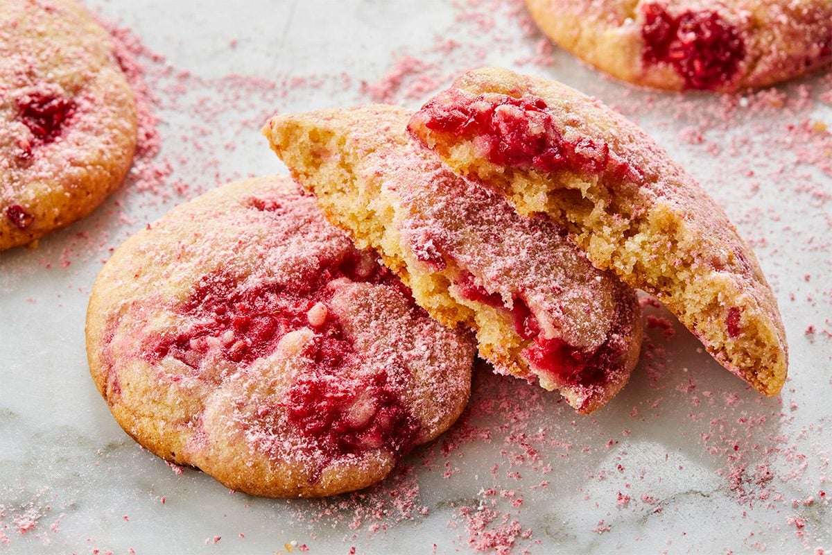 Two strawberry cookies on a white surface, one whole and one broken in half, revealing a soft, crumbly inside with visible strawberry pieces and a dusting of pink sugar on top and around them.