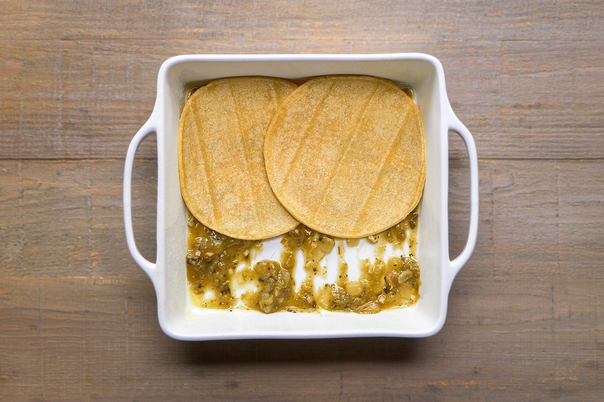A white square baking dish with handles contains two corn tortillas partially covering a layer of green sauce spread on the bottom, placed on a wooden surface.