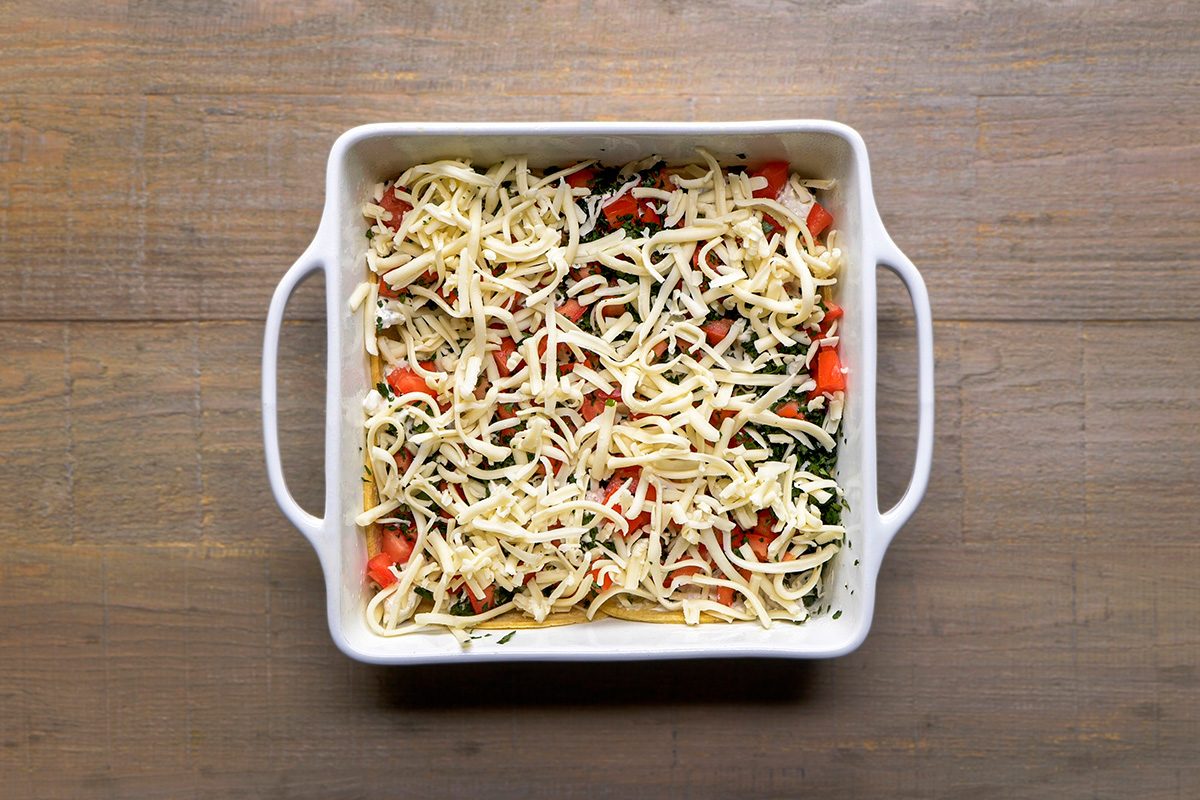 A white square baking dish filled with an uncooked casserole topped with shredded cheese, chopped red peppers, and green vegetables, placed on a wooden surface.
