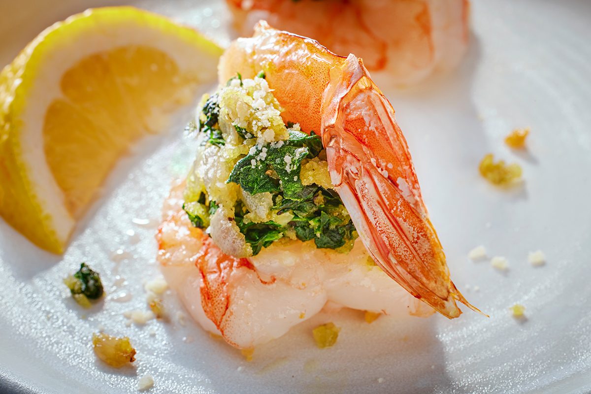 A cooked shrimp topped with herbs, breadcrumbs, and cheese sits on a white plate next to a lemon wedge. Some crumbs and herbs are scattered around the shrimp.