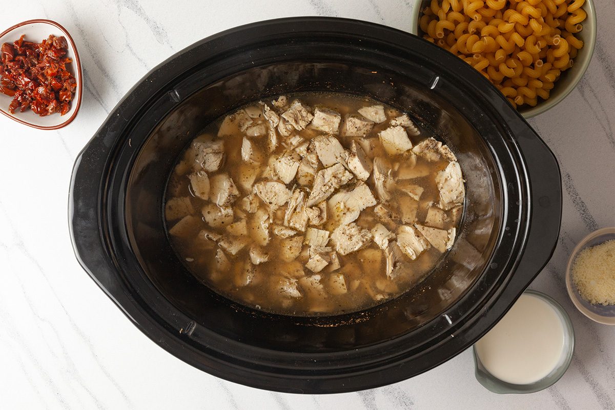 Overhead shot of a slow cooker filled with cooked, cubed chicken in broth, surrounded by small bowls of sun-dried tomatoes, elbow macaroni, grated cheese, and a bowl of milk on a white surface