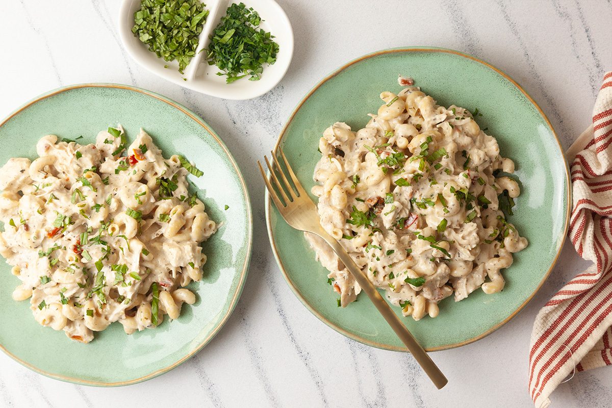 Overhead shot of two green plates of Slow-Cooker Chicken Boursin Pasta garnished with herbs on a white marble surface, with a gold fork on one plate, a small dish of herbs, and a red-striped napkin nearby