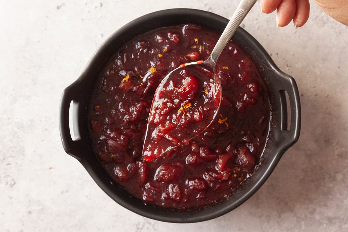 A hand holds a spoon scooping thick, chunky cranberry sauce from a black bowl on a light-colored surface.