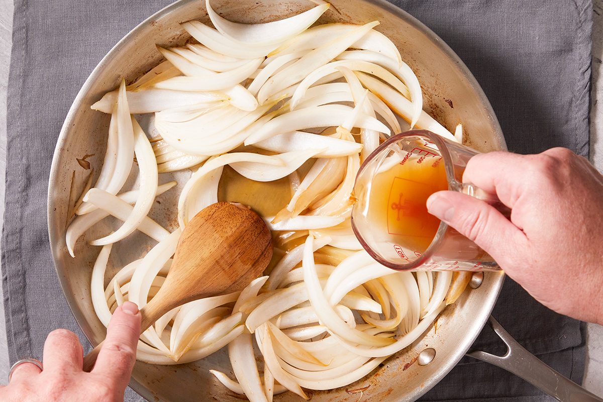 Sliced onions are being sautéed in a skillet; a person stirs with a wooden spoon while pouring liquid from a measuring cup over the onions.