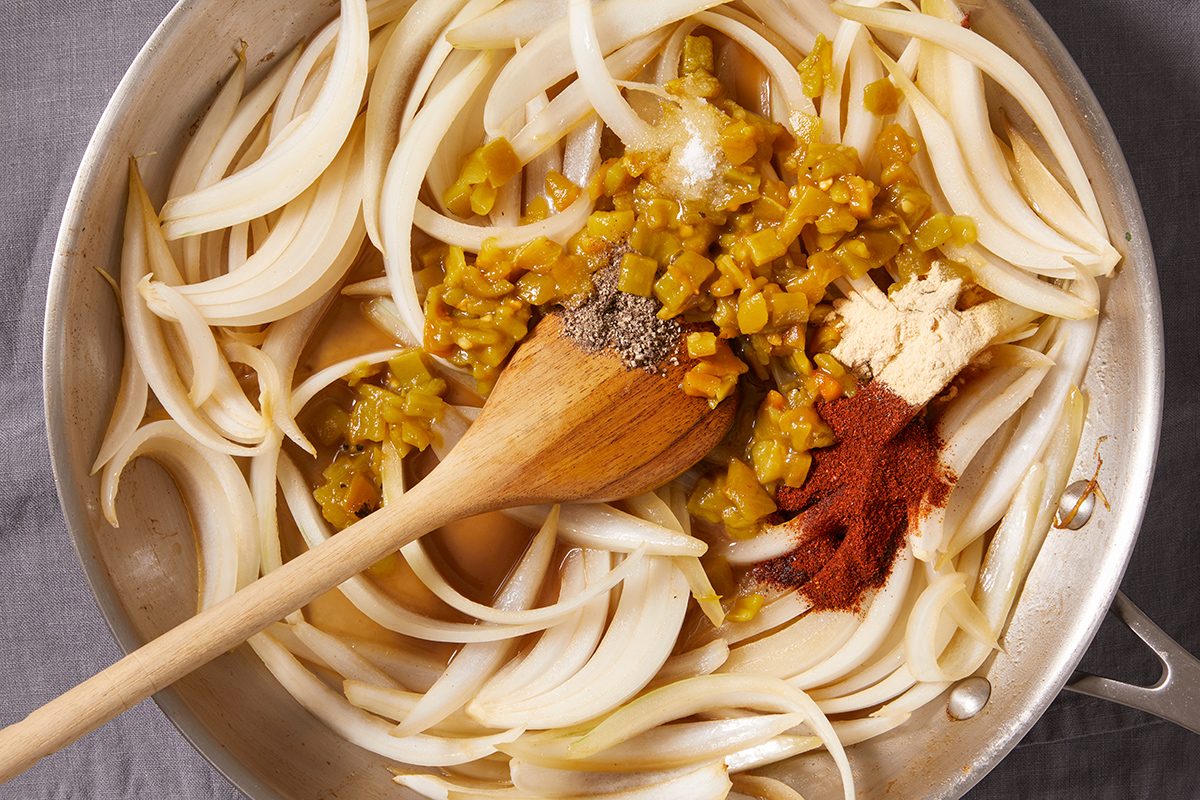 Sliced onions, diced green chiles, paprika, black pepper, and a dollop of minced garlic are being stirred in a pan with a wooden spoon.