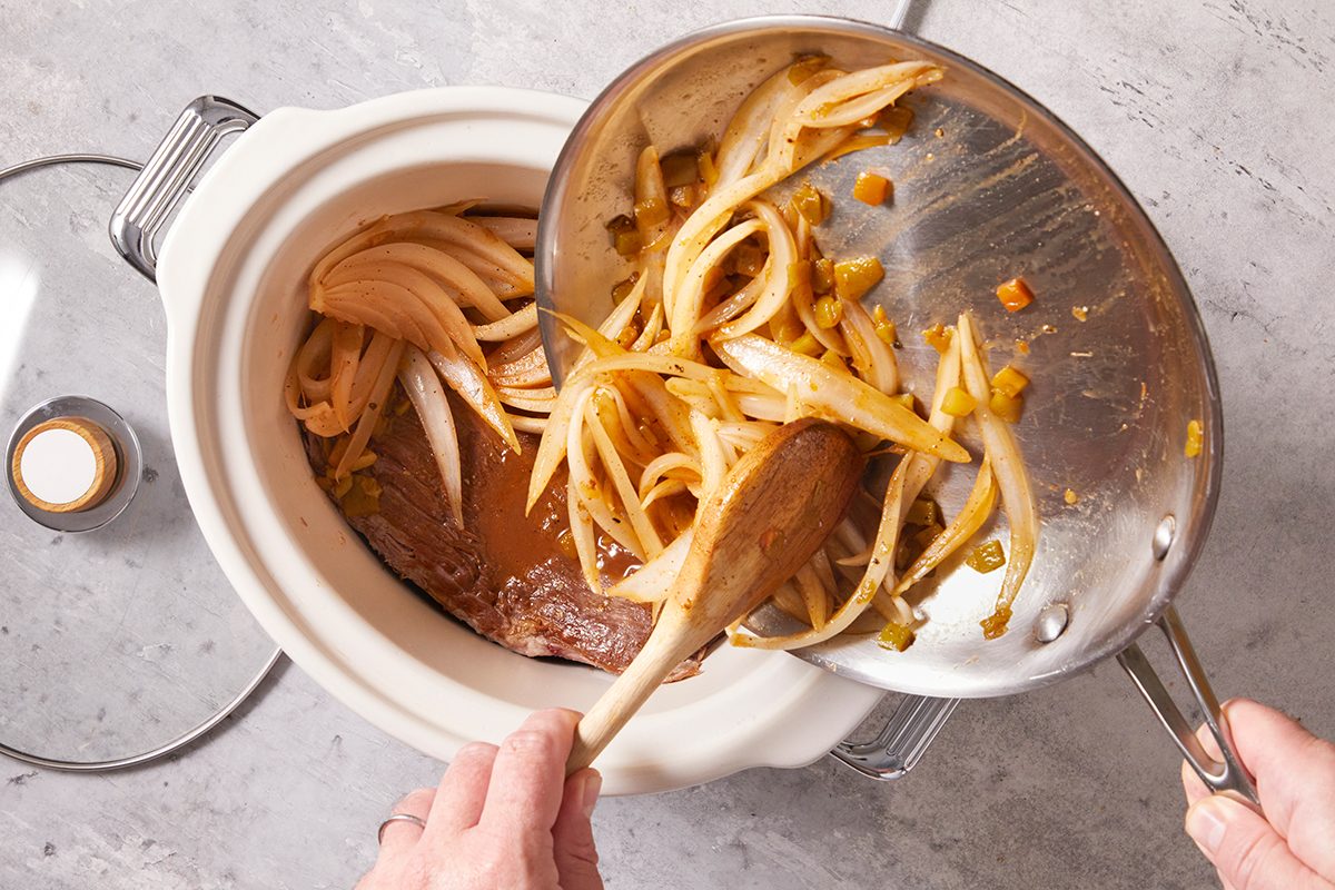 A person pours sautéed onions from a pan into a slow cooker containing a piece of meat in broth, preparing a meal on a gray countertop.
