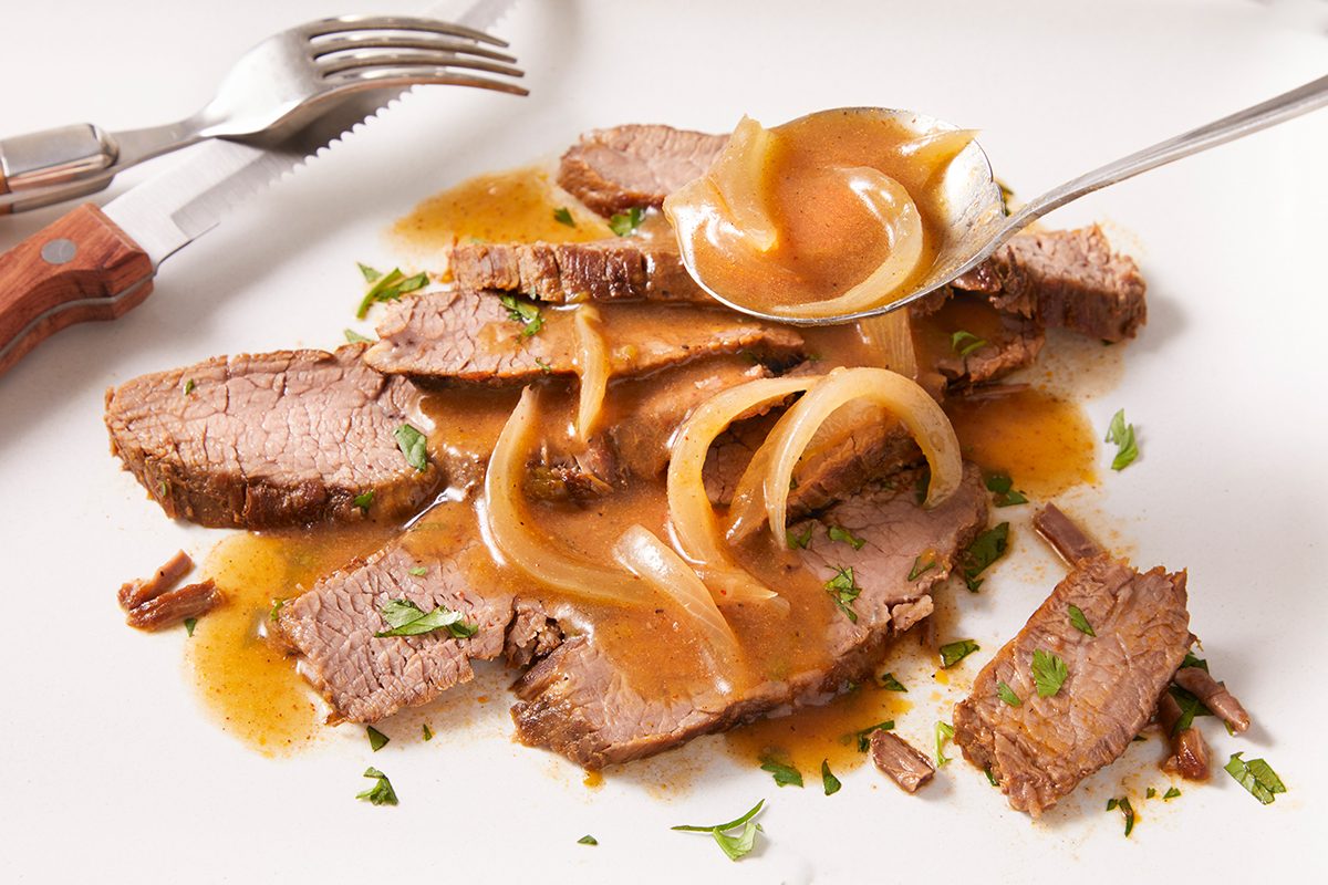 Sliced roast beef topped with brown gravy and cooked onions, served on a white plate. A spoon pours extra gravy over the meat, and a fork and knife rest nearby. Chopped herbs garnish the dish.
