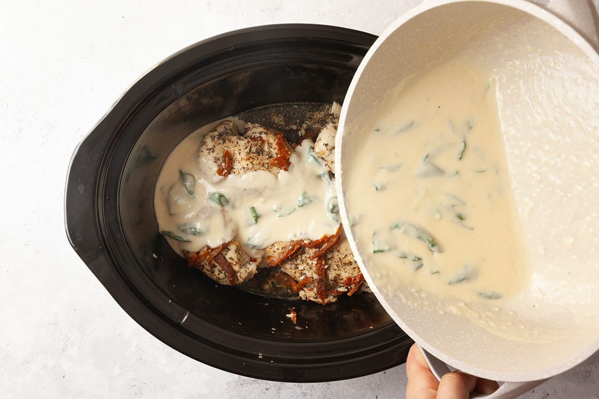 A hand pours a creamy white sauce with green vegetable pieces into a slow cooker filled with seasoned chicken pieces.