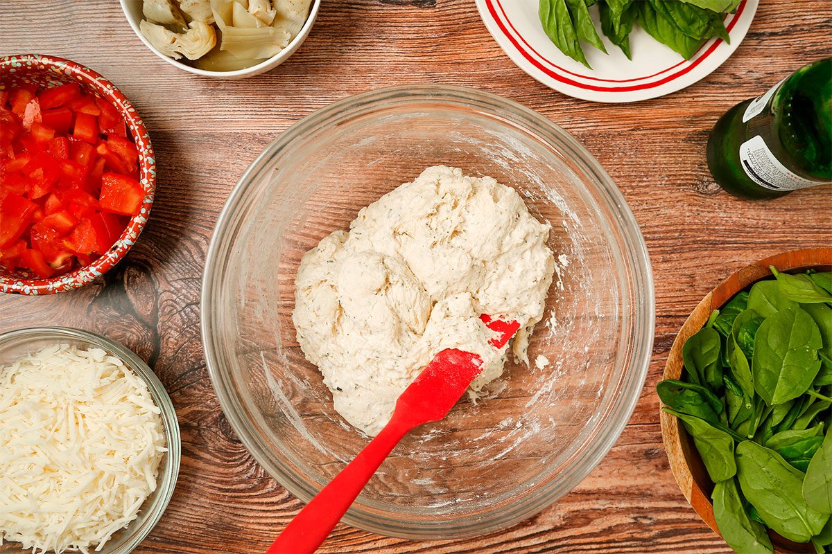 A glass bowl with pizza dough being mixed by a red spatula is surrounded by bowls of chopped tomatoes, shredded cheese, leafy greens, mushrooms, and fresh basil on a wooden surface.