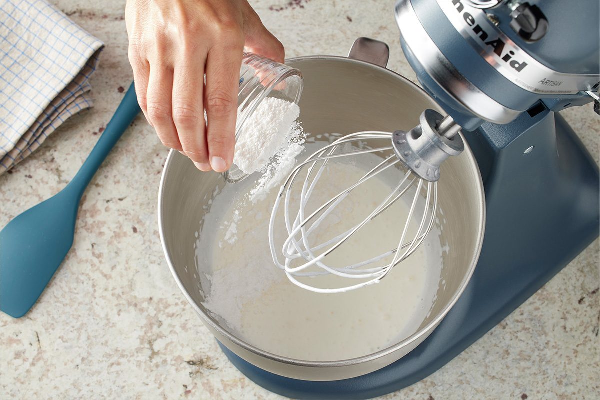 A hand pours a small glass bowl of white powder, likely sugar or flour, into the metal bowl of a stand mixer with a whisk attachment. A blue spatula and checked towel sit nearby on the countertop.