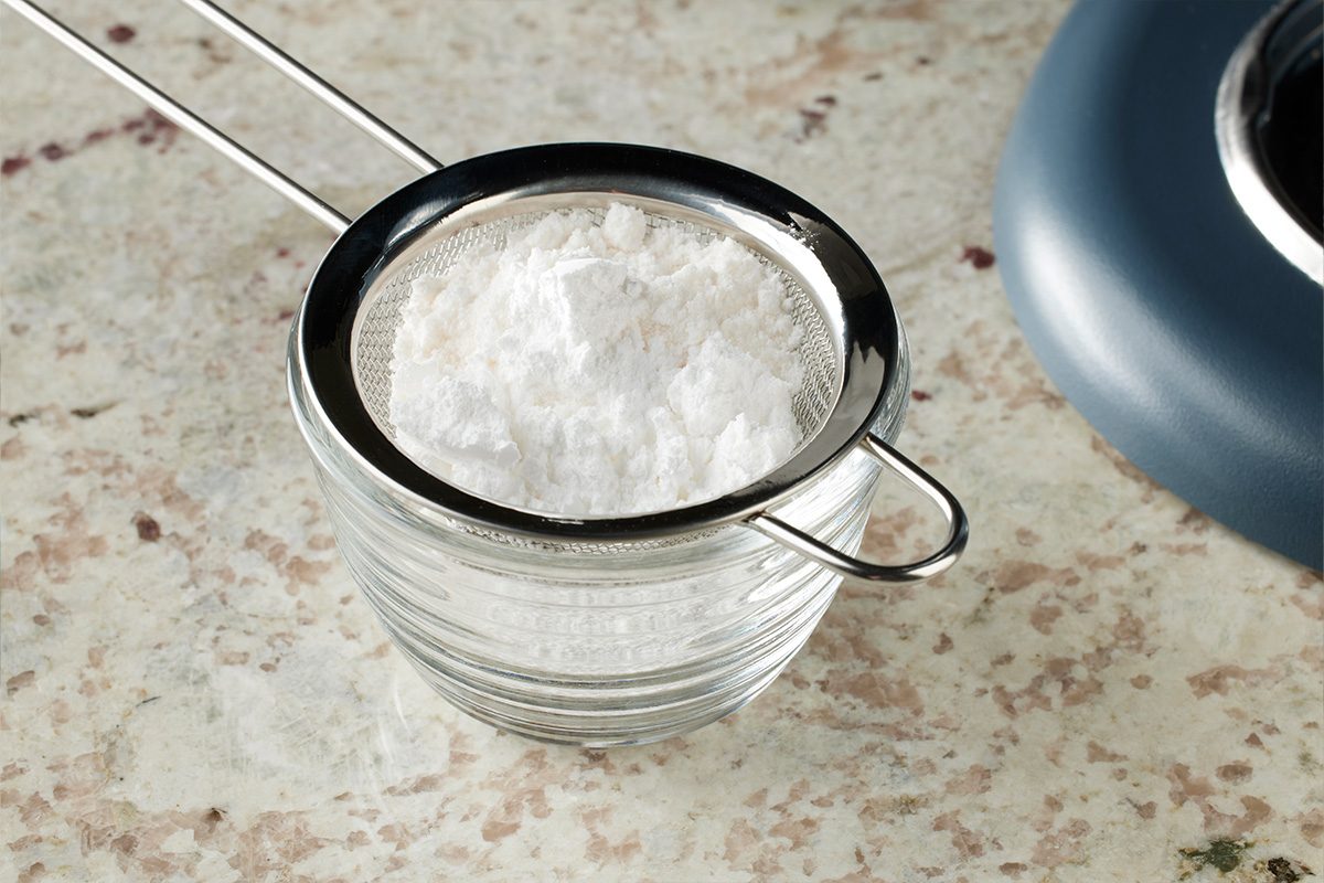 A metal sifter containing white powder, likely flour or powdered sugar, is placed over a clear glass bowl on a speckled countertop. A blue kitchen appliance is partially visible to the right.