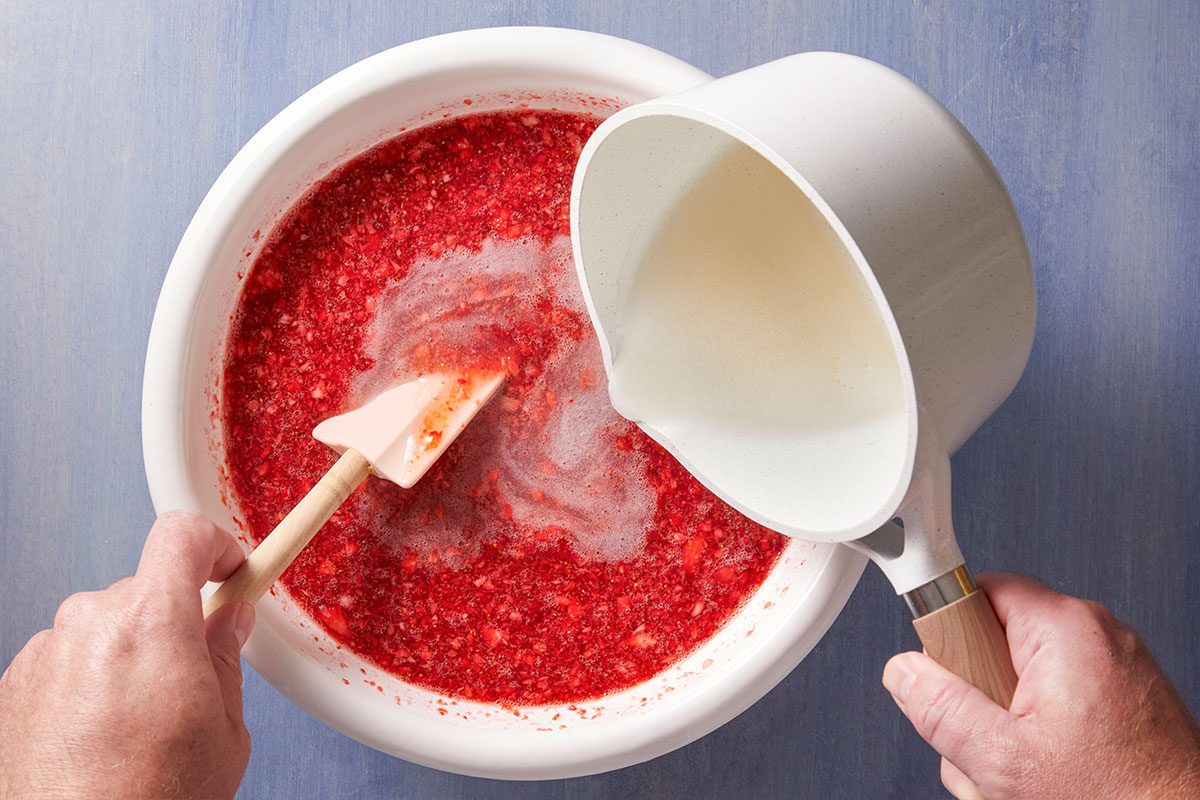 A person stirs a bowl of crushed strawberries while pouring a white liquid from a saucepan into the mixture, on a blue surface.