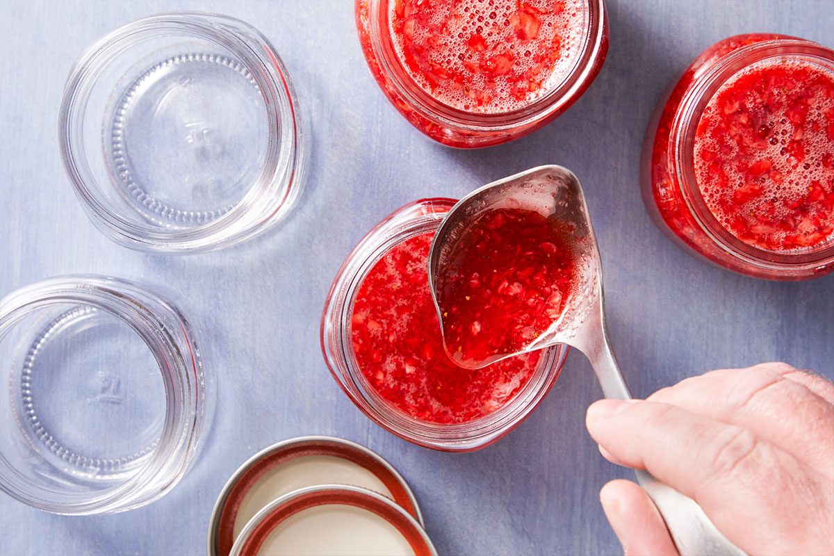 A hand uses a ladle to pour red strawberry jam into glass jars on a light blue surface, with jar lids and additional empty jars nearby.