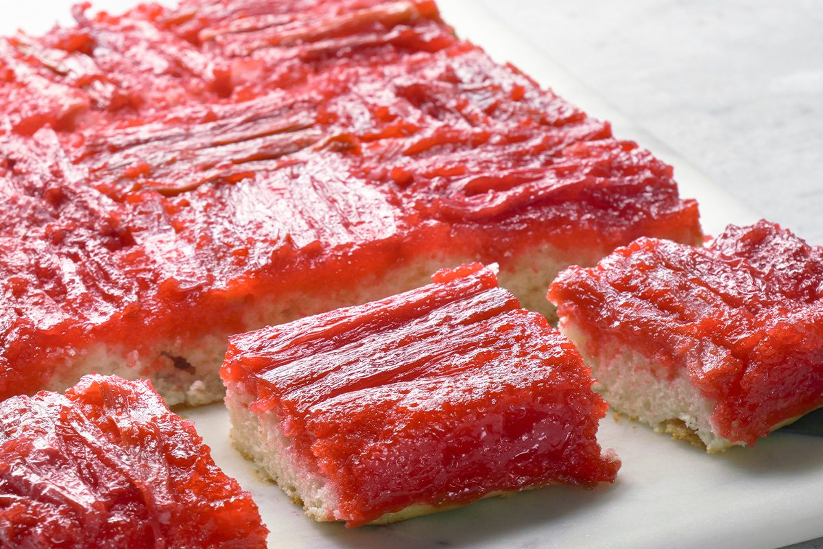 Several pieces of a rhubarb dessert bar with a shiny, pinkish-red rhubarb topping and a light-colored, cake-like base are arranged on a white surface. One piece is slightly separated from the others.