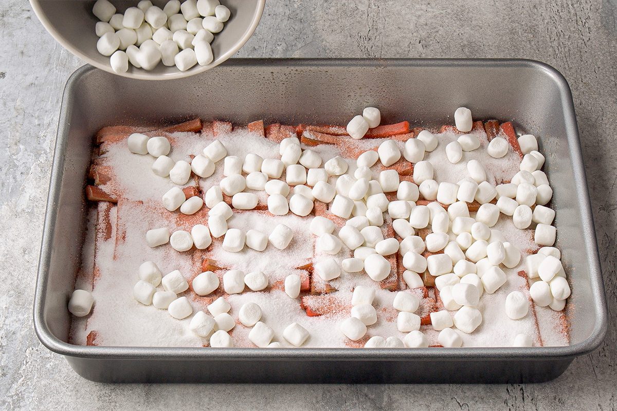 A rectangular baking dish filled with sliced sweet potatoes, covered in sugar and topped with mini marshmallows. A bowl is pouring more mini marshmallows over the dish.