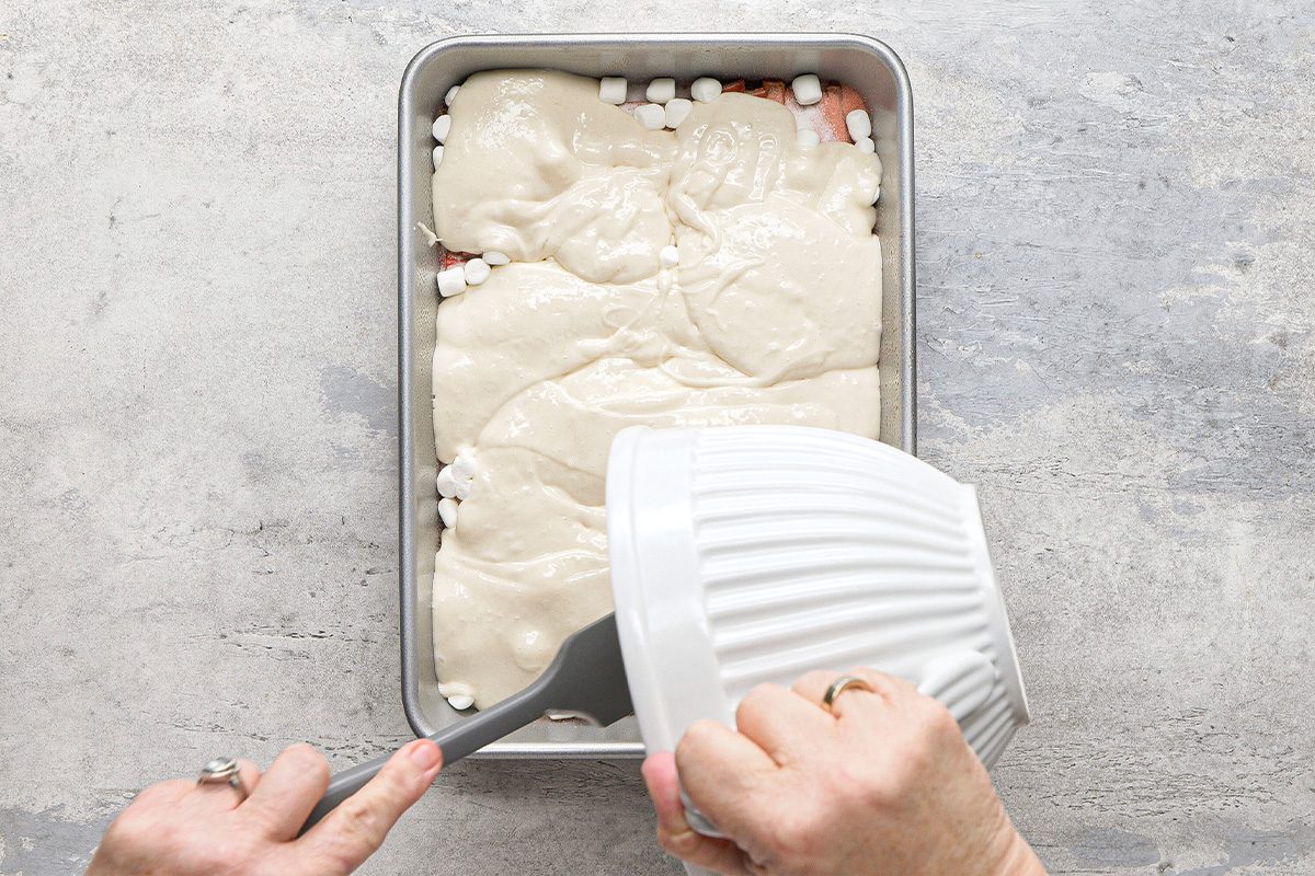 A person pours thick batter from a white bowl into a rectangular baking pan filled with mini marshmallows and other ingredients on a light gray countertop.