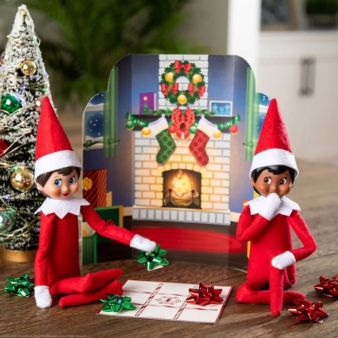 Two Elf on the Shelf dolls sit in front of a miniature Christmas fireplace scene, surrounded by wrapped presents, a decorated Christmas tree, and festive holiday decorations.