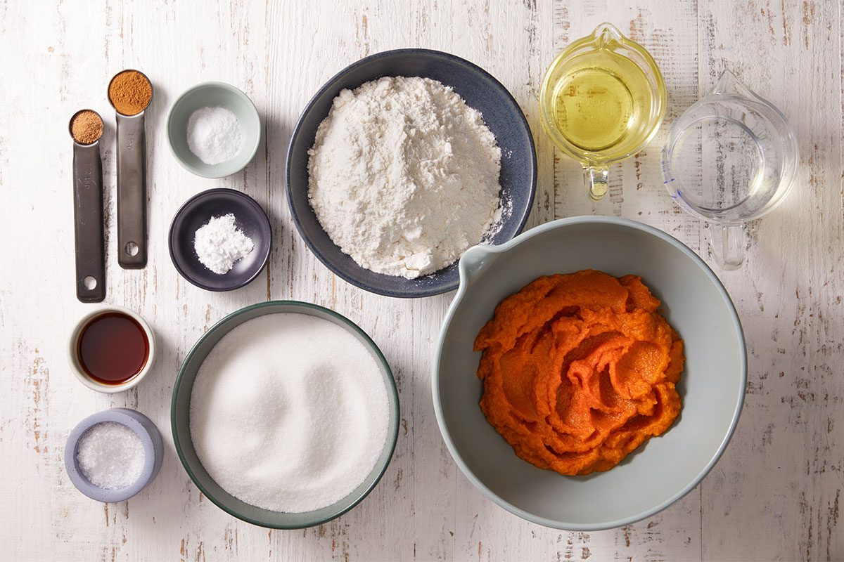 Baking ingredients arranged on a white surface, including a bowl of pumpkin puree, a bowl of flour, sugar, oil, water, and small bowls and spoons with spices, salt, baking powder, and vanilla extract.