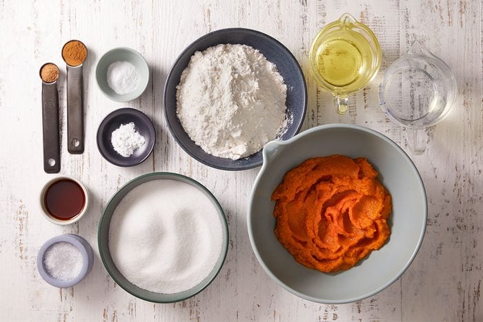 Baking ingredients arranged on a white surface, including a bowl of pumpkin puree, a bowl of flour, sugar, oil, water, and small bowls and spoons with spices, salt, baking powder, and vanilla extract.