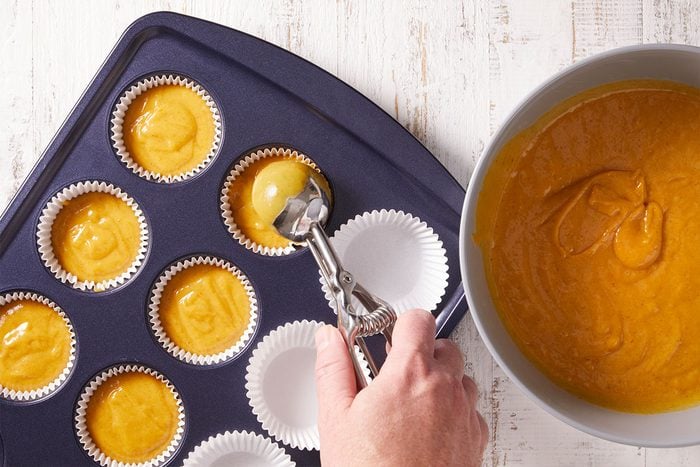 A hand uses an ice cream scoop to fill white paper cupcake liners in a muffin tin with orange batter from a nearby bowl on a white wooden surface.