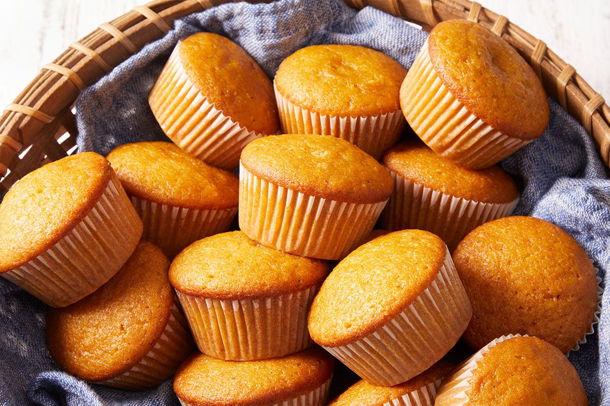 A basket filled with golden-brown muffins in paper liners, arranged on a blue cloth.