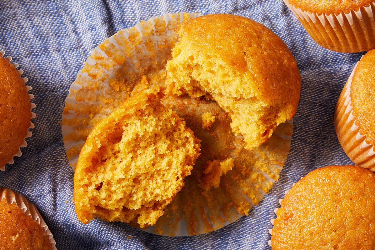 A split-open golden muffin sits in a paper liner on a blue cloth, surrounded by whole muffins, revealing its light, moist interior.