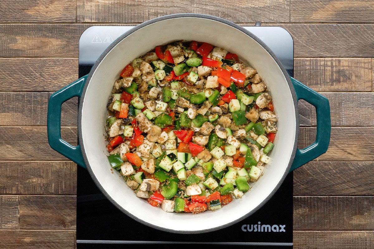 A white pot with green handles sits on a black stovetop, filled with chopped vegetables including red bell pepper, zucchini, and eggplant, all mixed and seasoned. The surface underneath is a wooden countertop.