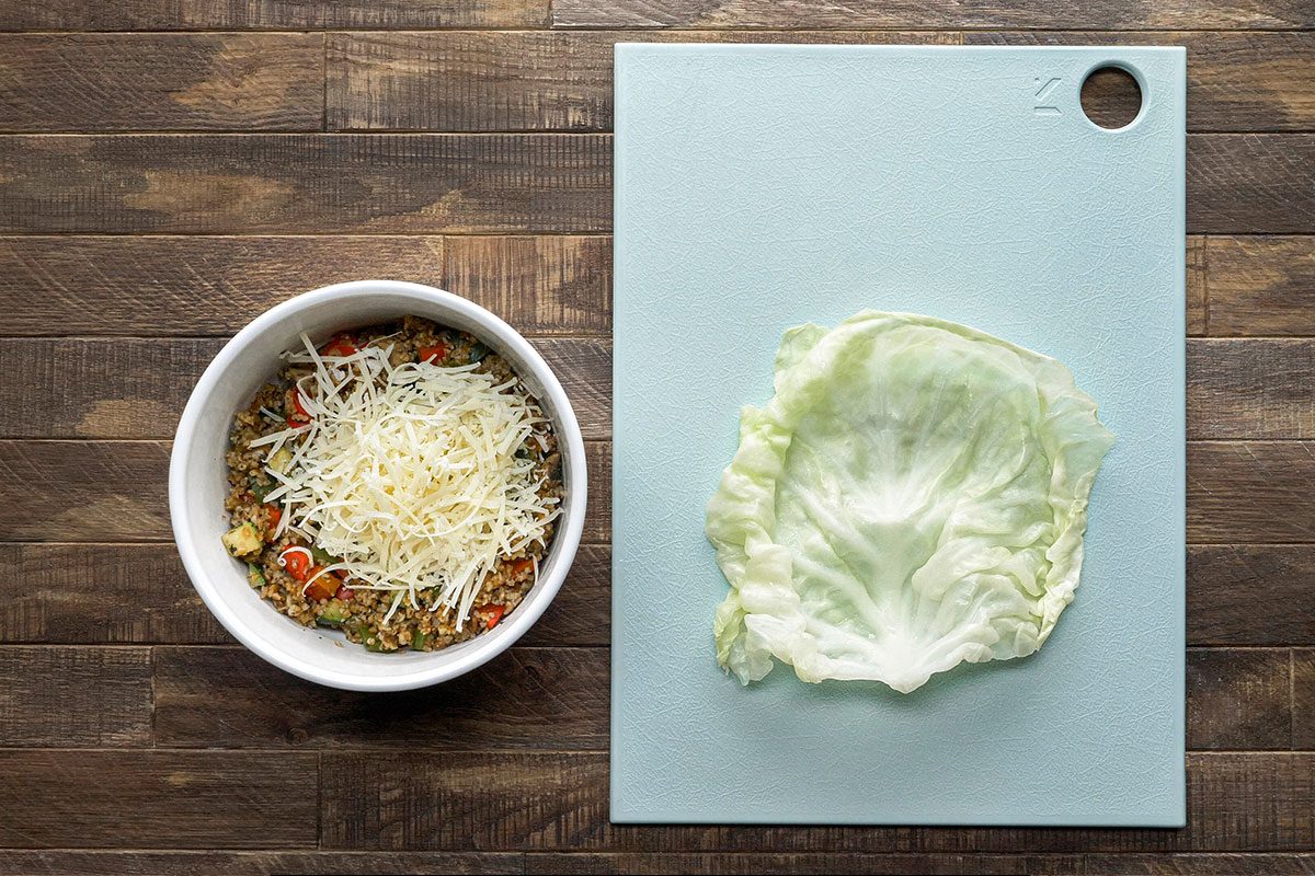 A bowl of mixed vegetables topped with shredded cheese sits next to a large iceberg lettuce leaf on a light blue cutting board, all on a wooden surface.