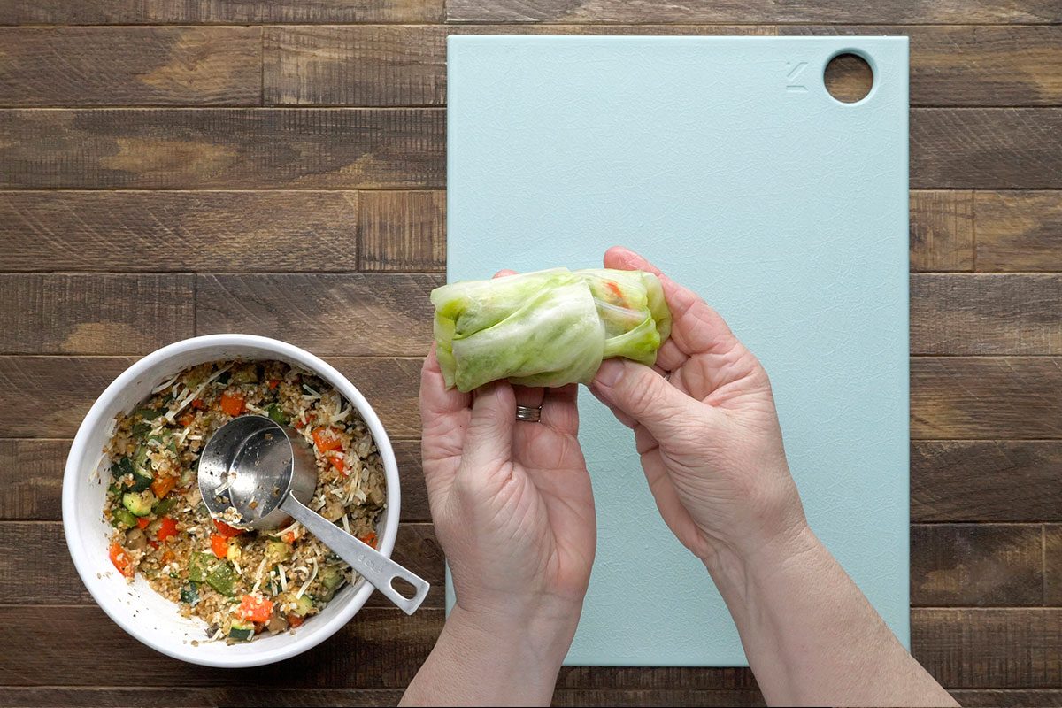 A person’s hands hold a wrapped cabbage roll above a white cutting board. Nearby is a bowl filled with a vegetable and rice mixture, with a spoon resting inside. The background is a wooden surface.