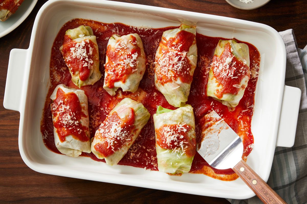 A white baking dish filled with stuffed cabbage rolls topped with tomato sauce and sprinkled with cheese, sitting on a dark wooden table with a spatula resting in the dish.