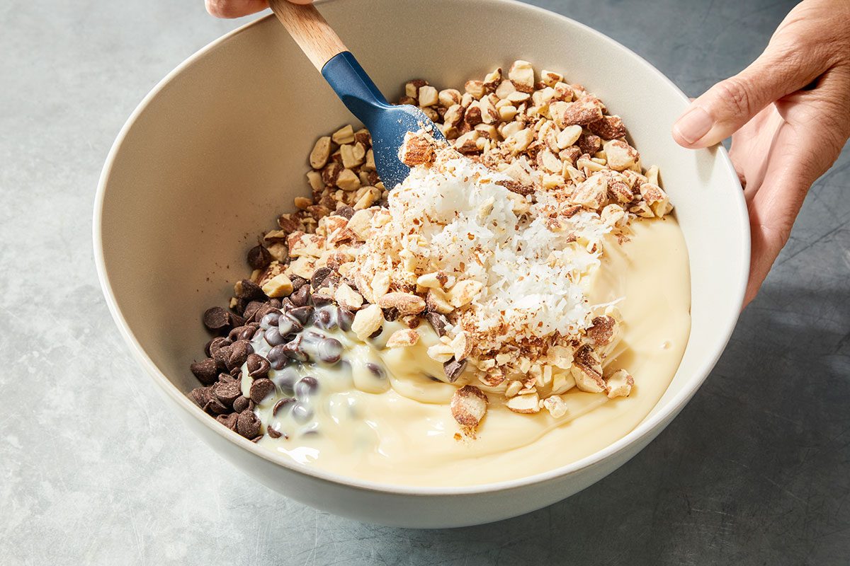 3/4 angle view shot of a bowl filled with sweetened condensed milk, chocolate chips, shredded coconut, and chopped nuts being mixed with a blue spatula by a person’s hand;