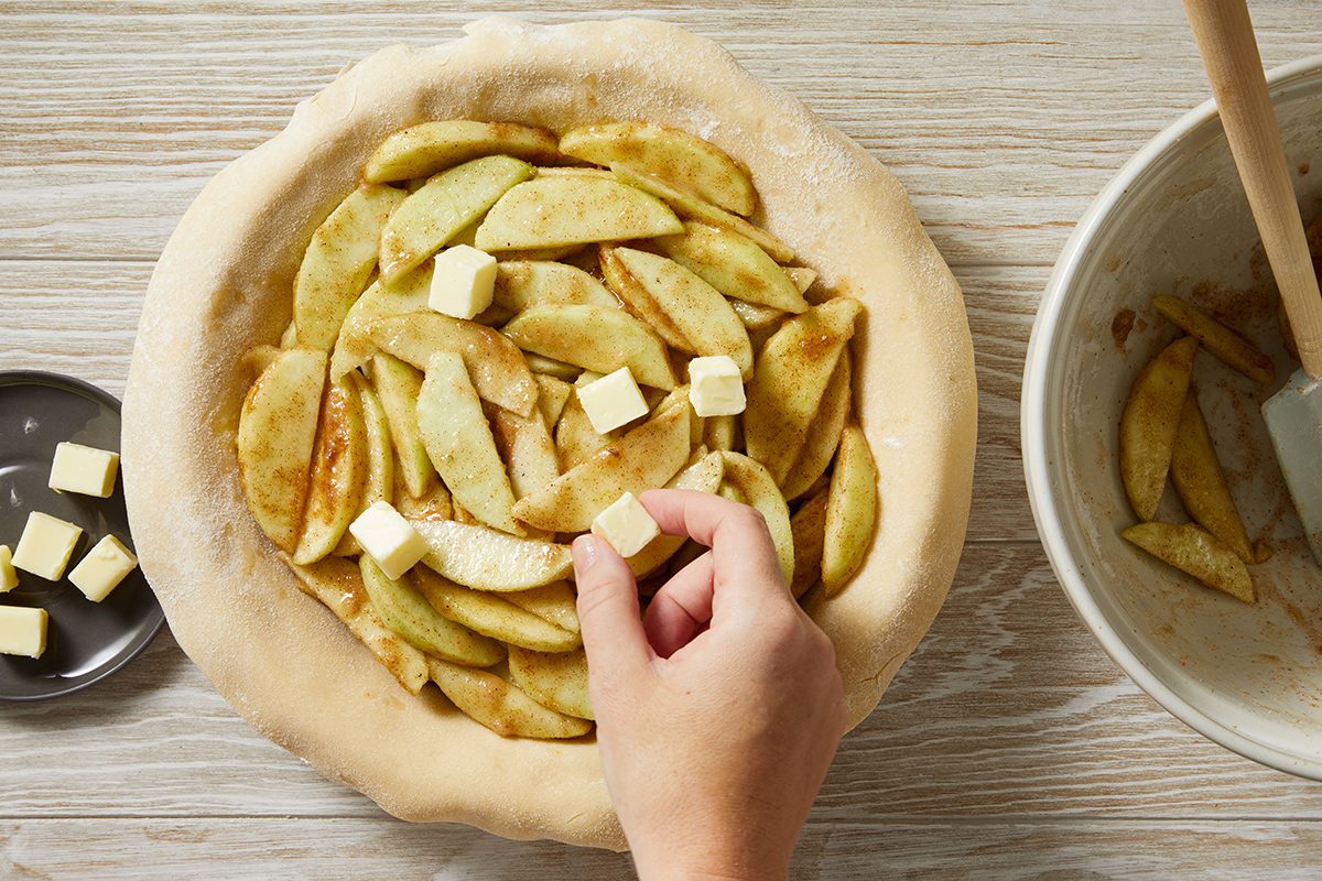 A hand places small cubes of butter on top of sliced apples arranged in an unbaked pie crust, with a bowl and spatula nearby on a wooden surface.