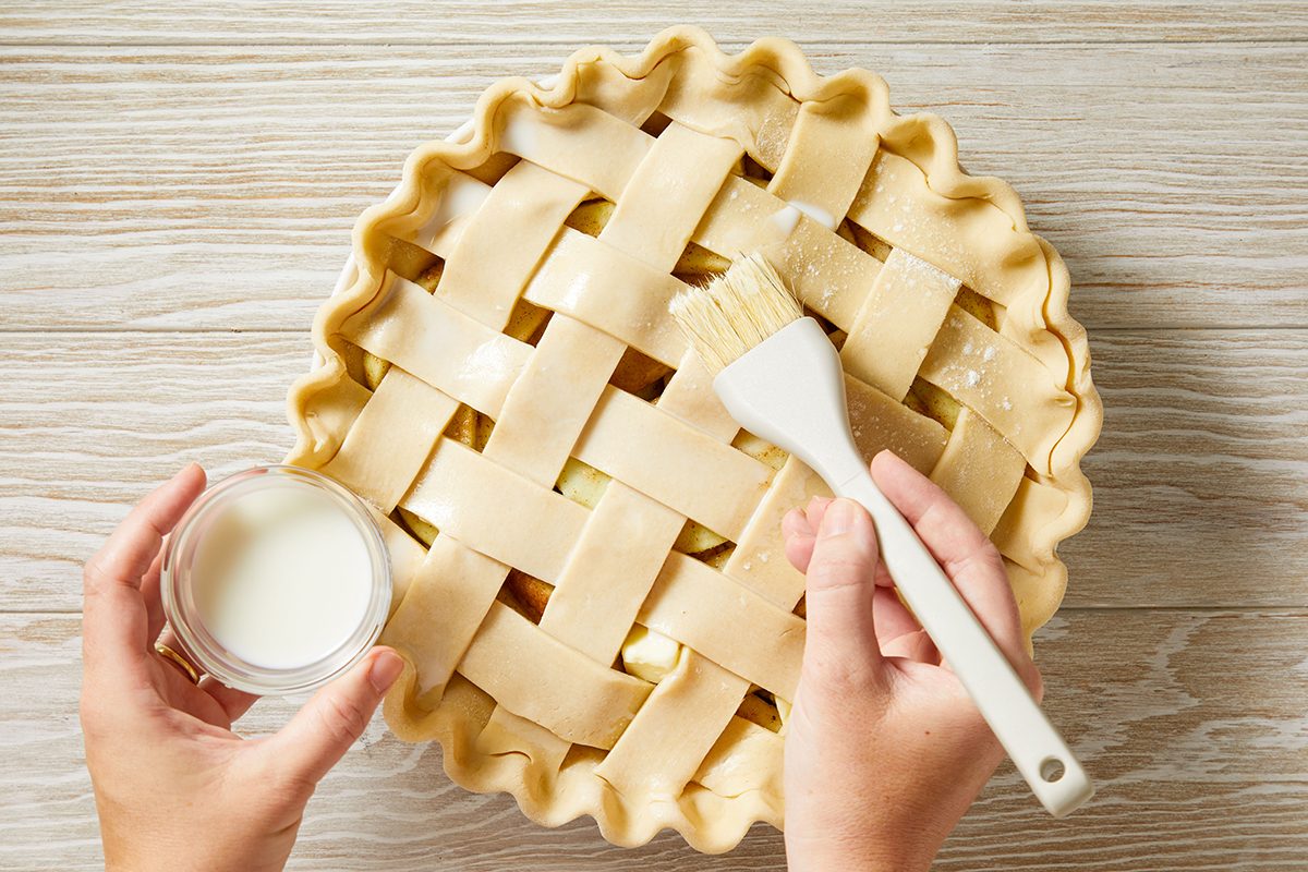 A person brushes egg wash onto a lattice-topped pie crust with one hand while holding a small bowl of egg wash with the other, on a light wooden surface.