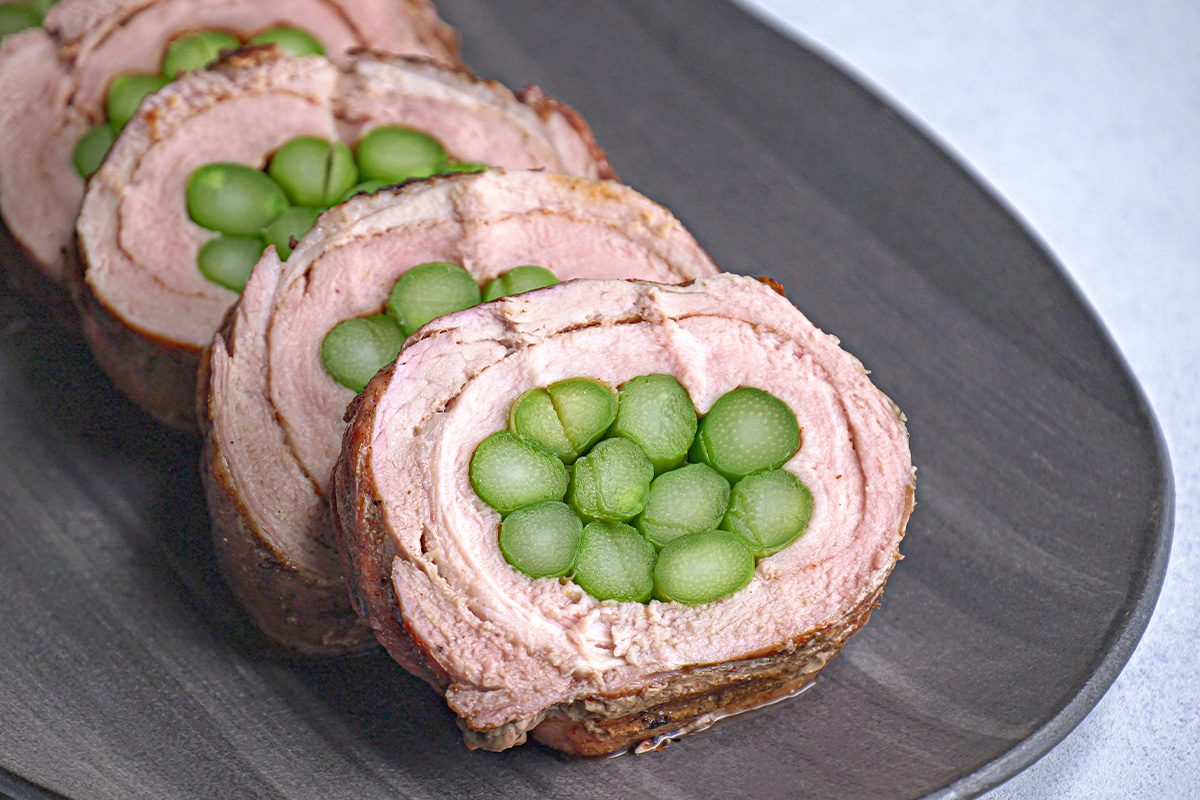 3/4th closeup shot of Asparagus Stuffed Pork Tenderloin slices rolled on a dark plate, each filled with green asparagus stems, neatly arranged for appeal