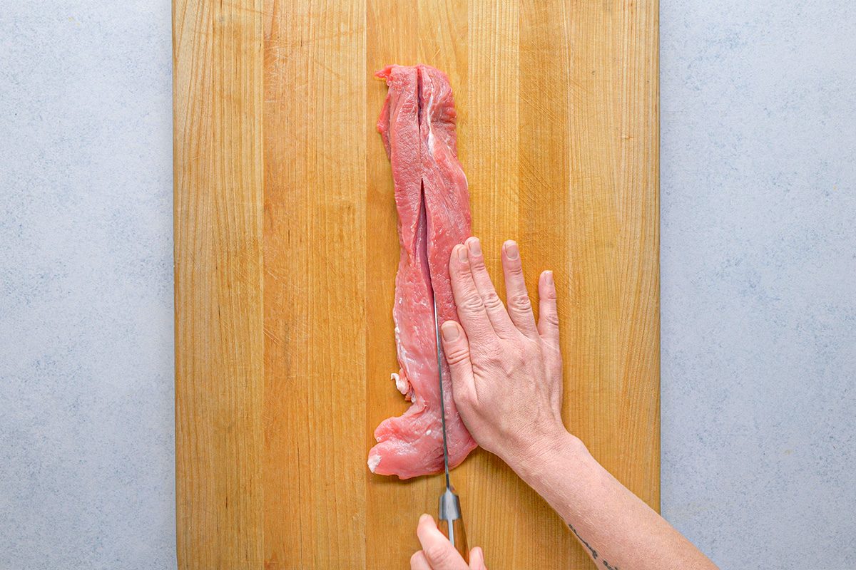 overhead shot of A person slices a raw pork tenderloin lengthwise on a wooden cutting board using a chef’s knife, with one hand holding the meat and the other guiding the knife