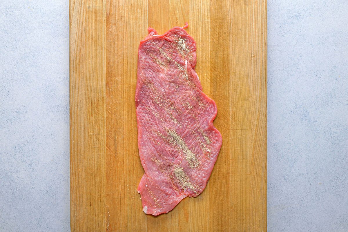 overhead shot of A raw, thin slice of meat lightly seasoned with pepper and salt lies on a wooden cutting board, against a light gray background