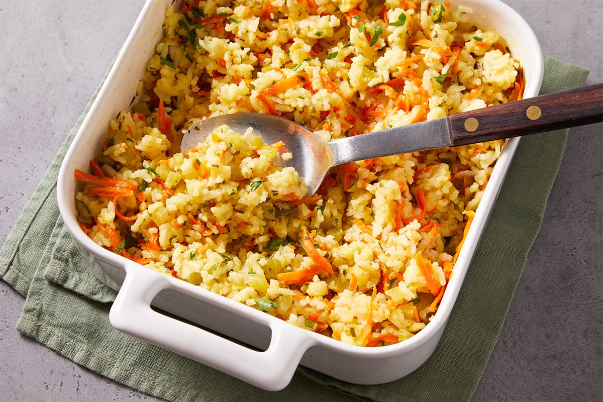 3/4 shot of baked rice pilaf with a serving spoon lifting a portion, revealing colorful bits of carrot and herbs.