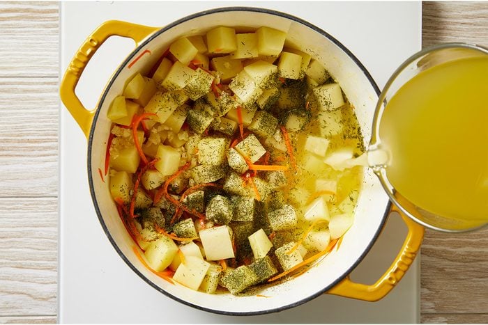 Overhead shot of a yellow pot filled with chopped potatoes, carrots, and herbs on a white surface, as broth is poured in to prepare a soup or stew;