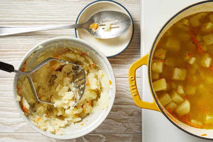 Overhead shot of a bowl of mashed potatoes with a masher, a plate with a fork, and a yellow pot of vegetable soup with visible potato chunks, all set on a light wooden surface;