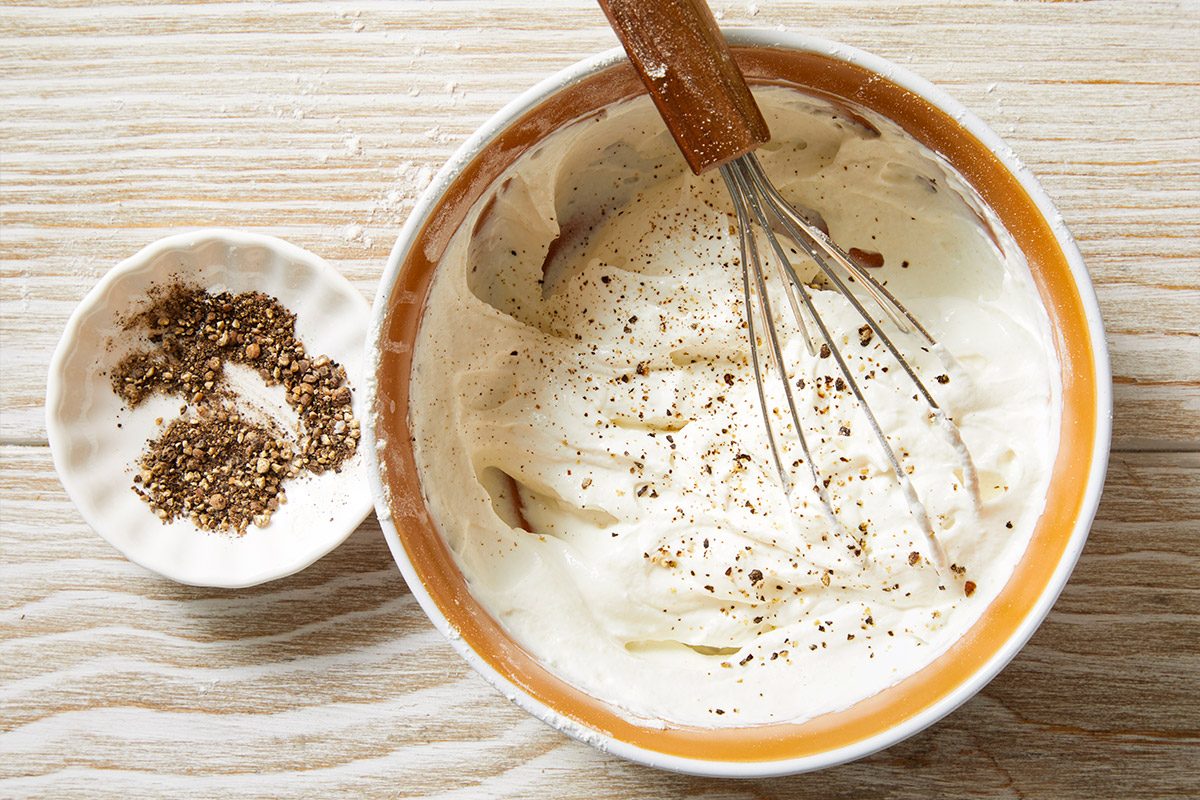 Overhead shot of a bowl of creamy white mixture topped with black pepper being whisked with a metal whisk, next to a small dish of ground black pepper on a light wooden surface;