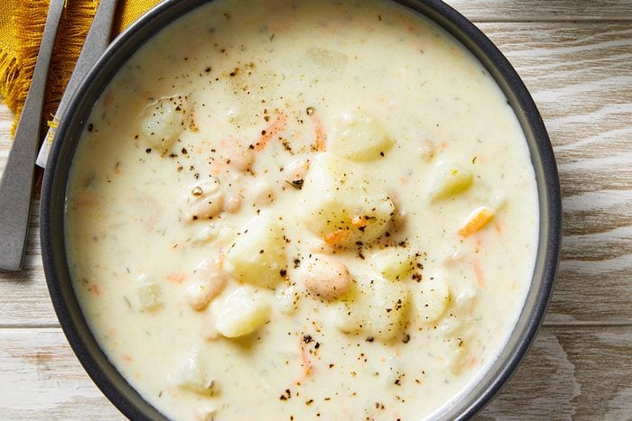 Top view shot of two bowls of Potato Soup with Beans topped with black pepper on a light wooden table, accompanied by a yellow napkin and two metal spoons;