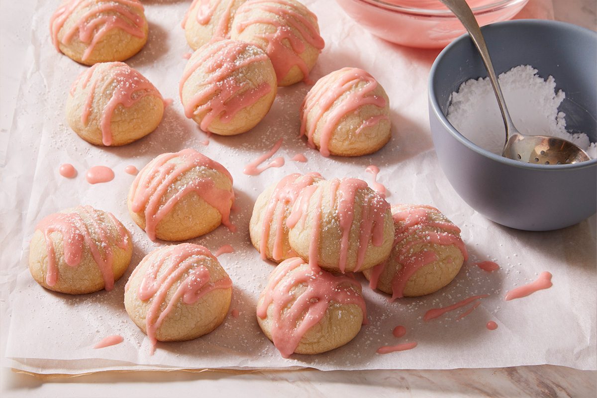 Overhead shot of a batch of Cherry Bonbon Cookies cooling on parchment with a small bowl of icing nearby.