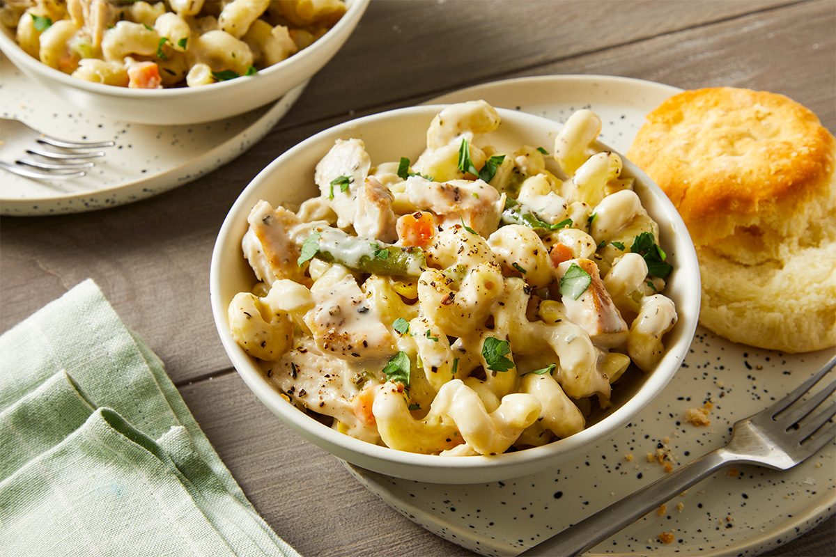3/4 angle view of a bowl of creamy chicken potpie pasta with curls of pasta, shredded chicken, and mixed vegetables, topped with black pepper and fresh herbs; a flaky biscuit sits on the plate beside the bowl, with a second serving visible in the background