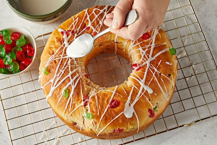 Drizzling glaze over baked bread with some candied fruit placed in a bowl on side.