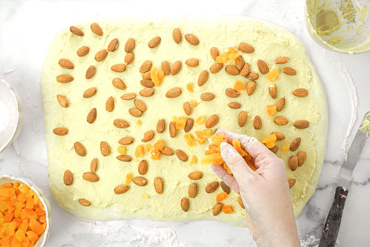 A hand sprinkles chopped dried apricots onto a rectangle of dough already topped with whole almonds, on a marble surface with bowls of ingredients nearby.