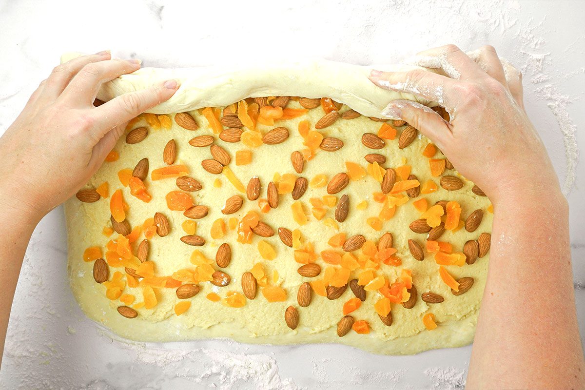 A person rolls up dough that is topped with chopped dried apricots and whole almonds on a floured surface.